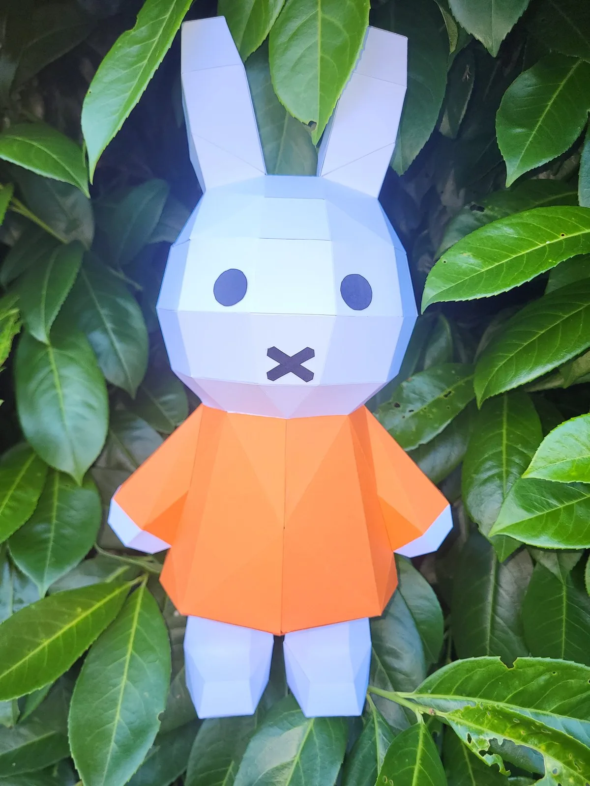 Lapin Miffy en papercraft blanc à robe orange devant un feuillage vert