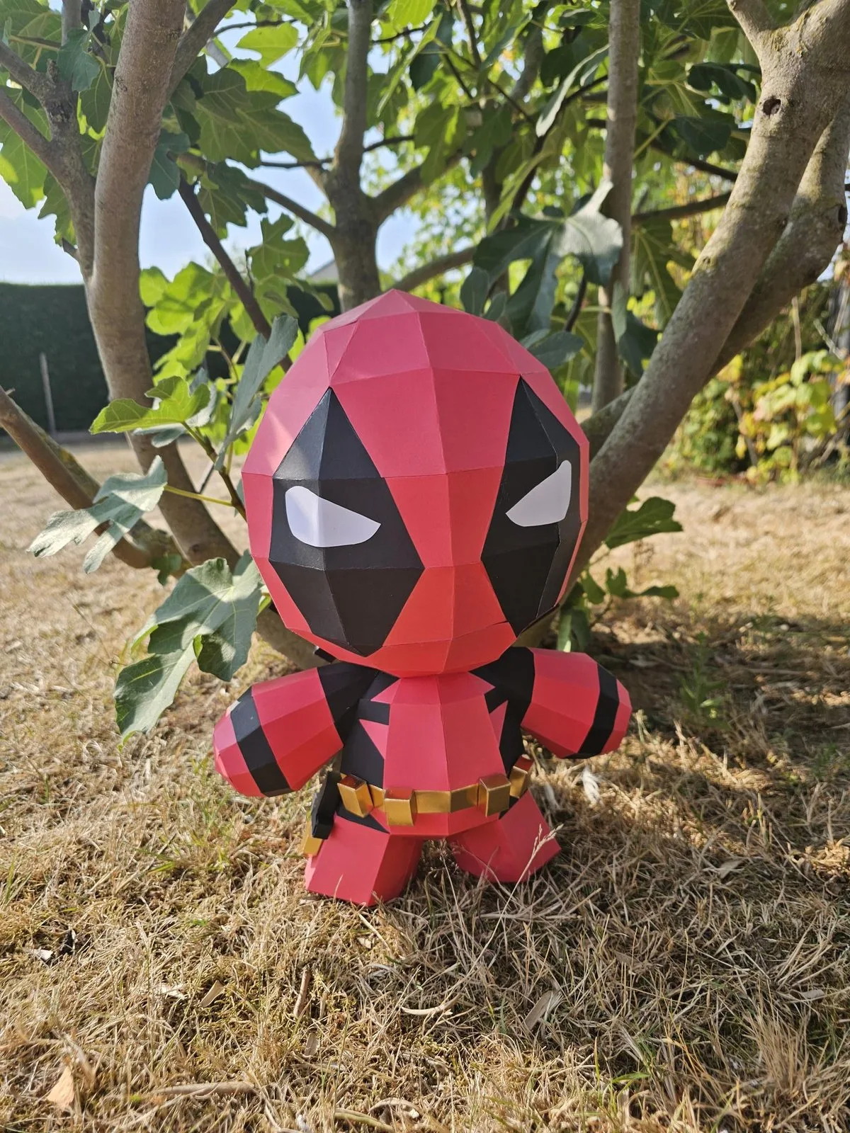Bébé Deadpool en papercraft rouge et noir posé au pied d'un arbre