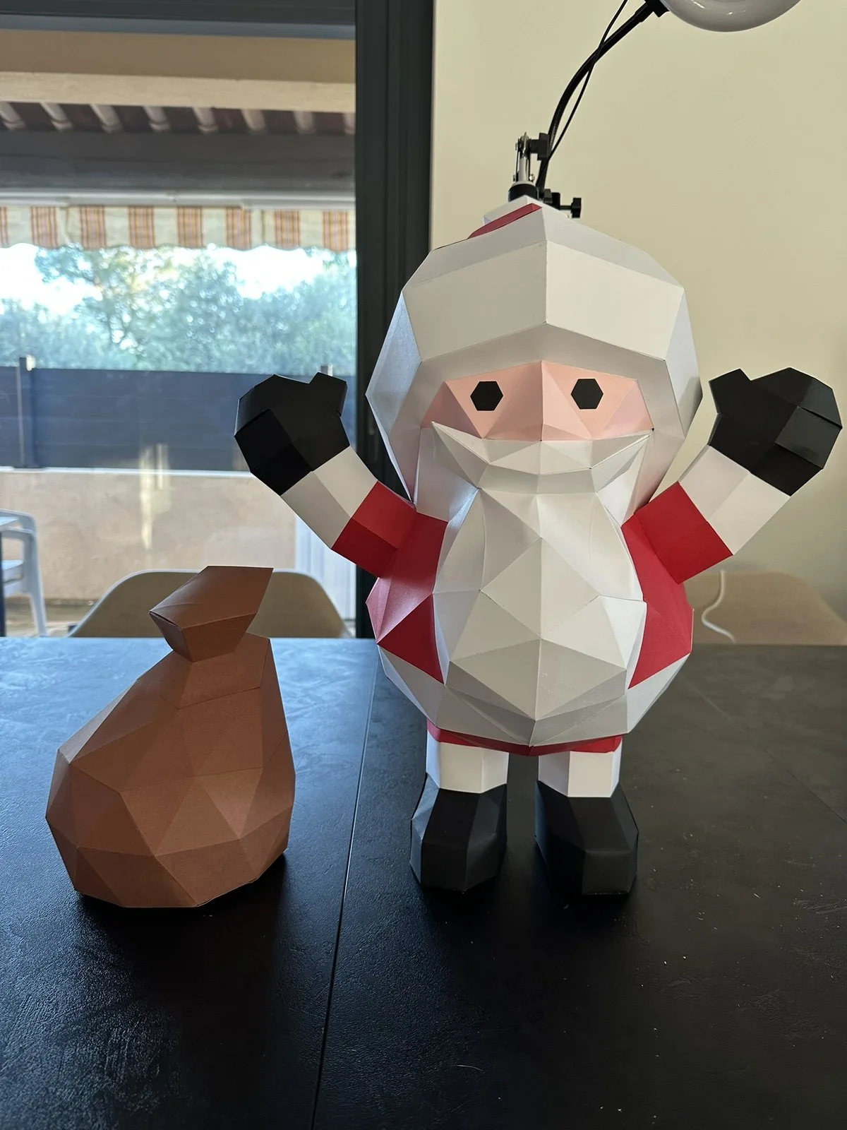 Père Noël debout bras levés moufles noires, avec hotte/sac marron à côté sur la table