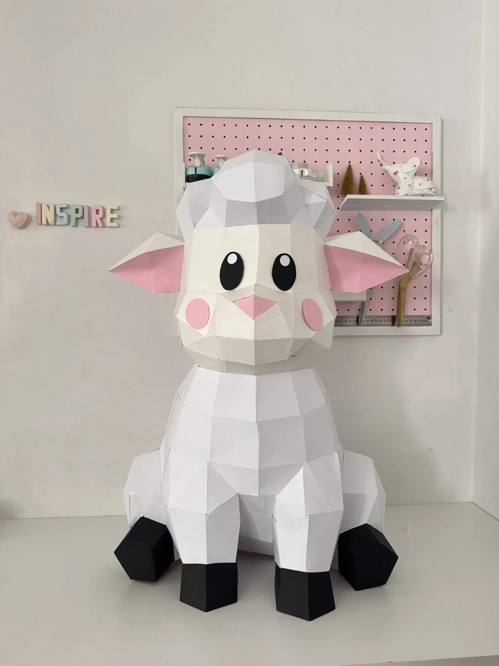 Mouton assis en papercraft 3D blanc, oreilles roses et sabots noirs, assemblé en papier et posé sur un bureau