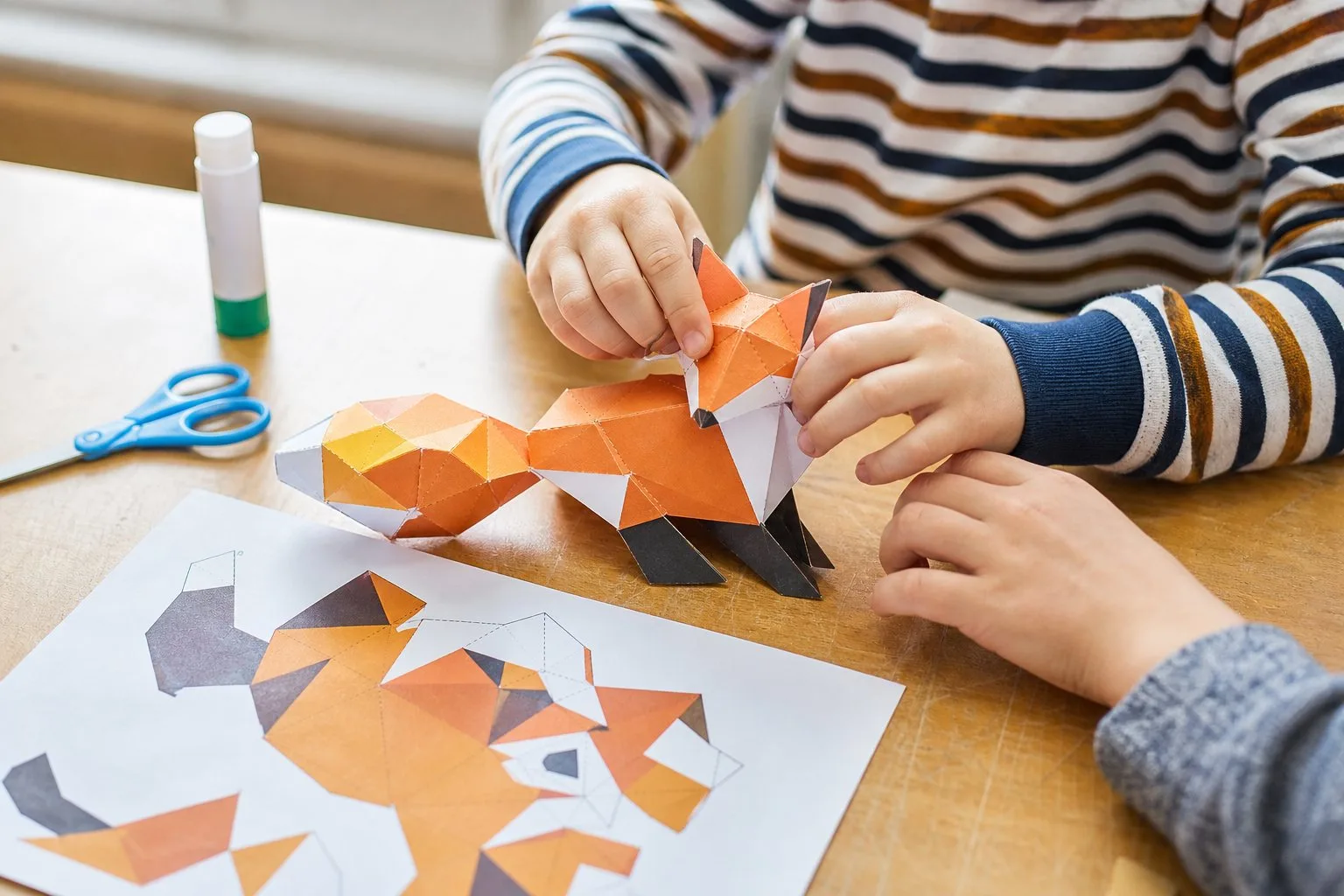 Handen van leerlingen die een low-poly papieren vos assembleren op een bureau
