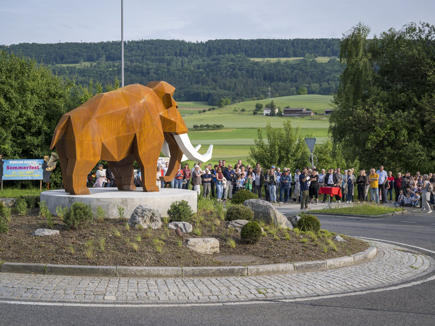 Mammouth monumental low-poly installé sur un rond-point, entouré du public lors de son inauguration